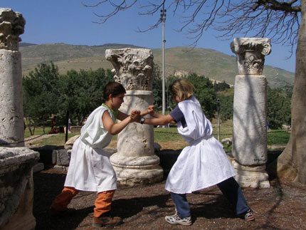 ילדים בתלבושות תקופתיות רומיות מדגימים דו-קרב, ברקע עמודים עתיקים עם כותרות קורינתיות